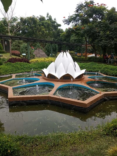 The image depicts an outdoor landscaped area, specifically a botanical garden or park in Ciherang, Indonesia, under an overcast sky. In the foreground, a large, ornate fountain structure is centered within a pond. The primary feature of the fountain is a multi-petaled, white lotus-shaped sculpture mounted on a star-shaped base. This base is brown with blue-lined hexagonal basins, each containing a water jet producing an upward spray. The surrounding pond water is dark and contains surface debris.

Beyond the fountain, the mid-ground consists of meticulously maintained garden beds featuring diverse foliage, including dense green groundcover, yellow shrubs, and red-leafed plants. A dark wooden pergola structure spans horizontally across the scene, supported by posts. Further back, a small, brown, pyramid-like structure is visible amidst the greenery. The background is dominated by lush, tall trees, some exhibiting sparse red blossoms.

On the far right background, two individuals are seated on a light-colored bench. One person wears a wide-brimmed hat and dark attire, while the other wears a hooded garment and lighter clothing. They appear to be resting or observing the surroundings. The overall environment suggests a public recreational or educational garden setting.