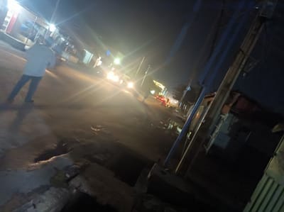 The image shows a dimly lit street scene at night in Narayi, Nigeria. A man in a light-colored shirt and dark pants is walking on the left side of the road. Several cars with headlights on are moving further down the street. Buildings line both sides of the street. Power lines and utility poles are visible on the right side of the frame. The foreground shows a rough, broken pavement. The overall light is low, with glare from the car headlights.