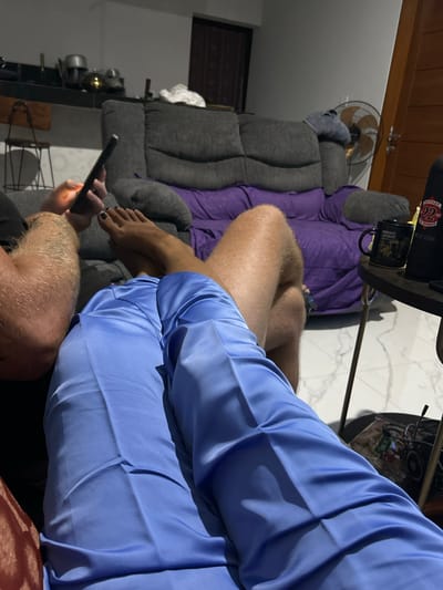 The media file shows a close-up view of people and objects in a room, likely in Sukawati, Indonesia. The scene is dimly lit, suggesting it might be evening or nighttime.
In the foreground, a person's legs are visible, covered by a light blue collared shirt. Their left leg is bent, and their bare foot, with dark nail polish, is resting on a gray couch. Another person's hairy legs are also visible in the lower part of the frame, suggesting two people are present.
A dark-colored smartphone is being held by a hand with dark nail polish, illuminated by its screen, indicating someone is actively using it. The phone is positioned between the two individuals.
The background features a plush gray sofa draped with a purple blanket. A black mug with an image and text is placed on a round table, along with other objects that are not clearly discernible. A ceiling fan is mounted on the wall. The floor appears to be tiled with a white and gray marble pattern.
On the table, a black object with a red and white logo is visible, with text that reads "SUPPORT 22MG" and "INDONESIA". The mug on the table has the text "NIGHT SAFARI" visible on it. The overall atmosphere appears relaxed and casual, with the individuals seemingly engaged in personal activities.