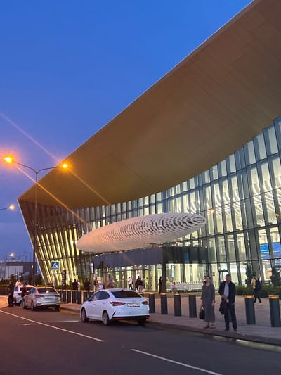 The image depicts the exterior of a modern airport terminal building at dusk. The building features a large, angled, golden-colored roof and a glass facade. A distinctive, elongated, white, sculptural element is suspended in front of the building. Several vehicles, including white sedans and a silver sedan, are parked on the roadway in front of the terminal. Pedestrians are visible near the entrance, some waiting or walking. Streetlights are illuminated, casting a warm glow. The sky is a deep blue, indicating twilight. The scene appears to be the entrance area of an airport, possibly serving the city of Vyazovka, Russia, given the car license plates with Russian characters.