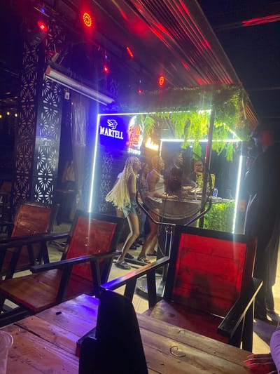 The image depicts an outdoor nighttime scene at what appears to be a bar or club in Howayi, Nigeria. The environment is lit by various artificial lights, including red and blue neon signs, and bright white LED strip lights. In the background, a neon sign displays "MARTELL Food House STUDIO." There is decorative greenery hanging above the illuminated signage. In the mid-ground, a large industrial fan is positioned prominently, with people dancing or standing behind it. To the left, decorative metal screens with floral patterns are visible on a column. The foreground features wooden tables and chairs, with some of the chairs having red wooden seats and black frames. A person's hand with long fingernails is visible in the lower right corner, suggesting they are seated at one of the tables. The overall atmosphere is lively, characteristic of a nighttime entertainment venue.