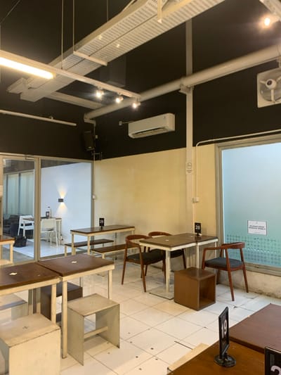 The image shows the interior of a cafe in Malang, Indonesia, located at latitude -7.941 and longitude 112.608. There are several tables with white metal frames and wooden tops, arranged in a square formation. The tables are surrounded by white metal chairs with wooden seats, except for one with a dark brown chair. The cafe features a white ceiling with exposed white pipes and a white air conditioner mounted on a black wall. The cafe is empty, with no people present. The walls are painted in yellow and black tones. Two windows are visible. The window on the left side looks into another room with a couch and another table with chairs. The window on the right side is frosted. The table in the foreground has a sign with the number "19" on it. There is a sign on the wall beside the window on the right with an inscription in Indonesian. The floor is tiled white.