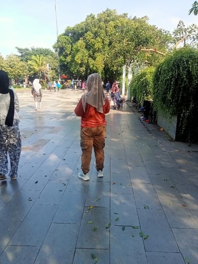 The image shows a wide, paved walkway, likely a pedestrian thoroughfare, with a woman wearing a long, brown dress, a long-sleeved orange shirt, and a beige headscarf, standing facing the viewer in the center of the frame. She is walking in a park, in the city of Pati, Indonesia, located at coordinates -6.752, 111.04. The walkway is made of smooth, gray tiles, and there is a large tree on the left side of the frame. There are some people in the background walking away from the viewer and the woman, indicating a park or public area. The foliage is green and lush. There is a small amount of litter on the ground. The woman is the only person in the image who is standing still, suggesting she is the subject of the photo. The image has a bright, natural lighting, suggesting it was taken during the day.  The picture appears to have been taken during a typical day in a public park.