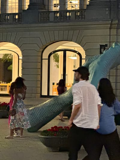 In the city of Amsterdam, Netherlands, at dusk, a group of people are gathered near a building with arched windows and classical architectural details. A large, teal-colored sculpture, possibly an abstract representation of a hand or limb, dominates the foreground, adorned with intricate text.
Several individuals are visible:
* A woman in a floral dress, her hair dark and pulled back, stands on the left, facing away from the camera. She is wearing flat shoes and carrying a small crossbody bag.
* To her right, a man wearing a white, long-sleeved shirt, dark pants, and a black baseball cap is seen from the back. He appears to be looking towards the sculpture.
* A woman with long, dark hair, wearing a blue top, stands to the right of the man in the white shirt, also seen from the back.
* Further back and to the left, another woman with dark hair, wearing a maroon top and a dress, is partially visible.
* Another woman in a pink dress is on the far left of the frame, facing left.
The environment is paved, suggesting a public square or walkway. Planters filled with red flowers are strategically placed around the base of the sculpture, adding a touch of color to the scene. The building in the background is illuminated from within, casting a warm glow through the windows and highlighting the building's stonework and balconies. The overall atmosphere is one of casual evening gathering, with people interacting or observing the public art installation.