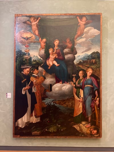 This is a painting, likely an altarpiece, depicting a religious scene with multiple figures in a landscape. In the foreground, two male figures are shown, one wearing a black Dominican habit and the other in ornate, layered clothing. The figure in the habit appears to be holding a book and possibly a palm frond, while the other figure has a halo and is holding a sword and a chalice. Behind them, a group of female figures, including a woman with a halo, and children, are gathered in the upper part of the painting, with cherubic figures floating around them. The background features a rocky landscape with a body of water and trees. The overall impression is one of solemnity and reverence. The painting is framed by a simple wooden border. A small label is affixed to the wall below the painting. There are no clear indicators of time of day or weather. The context of Palermo, Italy suggests this is likely a Renaissance or Baroque religious artwork.