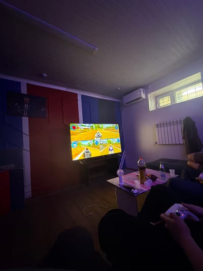 The image depicts an indoor setting, likely a living room or entertainment area, with a group of people engaged in playing video games. A large television screen displays a racing game with cartoon-like graphics. In the foreground, one person's hands are visible holding a white video game controller, suggesting active participation. A small table in front of the television holds various items including drinks, cups, and a small fan. The room is dimly lit, with ambient lighting casting purple hues on the walls. The walls themselves are painted in distinct vertical sections of blue, red, and white. An air conditioning unit is mounted high on one wall, and a radiator is visible beneath a window. Another smaller screen is mounted on the wall above and to the left of the main television. The flooring appears to be wooden. The overall impression is of a casual, relaxed atmosphere focused on gaming. There is no discernible information within the image to suggest a location in Ayteke Bi, Kazakhstan.