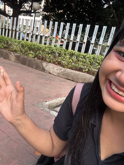 A woman smiles at the camera in front of a metal fence and lush greenery. She is wearing a black shirt and a pink backpack. Her right hand is held up, showing a small white object between her thumb and index finger. The ground is paved with red bricks. The scene appears to be an outdoor public space in West Jakarta, Indonesia.