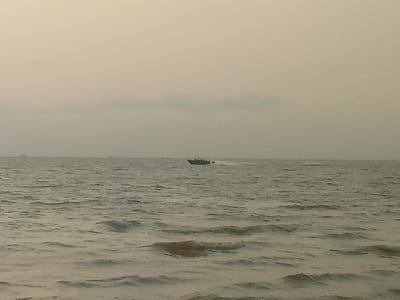 A wide expanse of water dominates the scene under a muted sky. Waves ripple across the surface, reflecting the gray tones of the sky. A small boat, appearing relatively small in the distance, cuts through the water, leaving a white wake behind it. Distant shapes, possibly ships or land, are barely visible near the horizon, adding depth to the perspective. The overall tone is somber and subdued, emphasizing the vastness of the sea.
