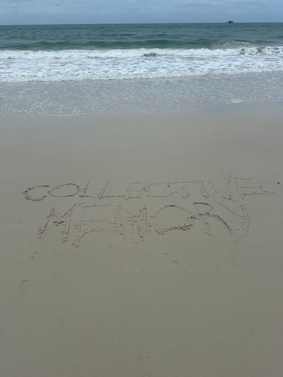 The image depicts a beach scene in Ao Hin Khok, Thailand. The sand in the foreground has the words "COLLECTIVE MEMORY" written in it. Gentle waves are breaking on the shore, and the ocean stretches out to the horizon under a cloudy sky. A small boat is visible in the distance on the water. The overall scene is tranquil and captures a moment of inscription on the beach.