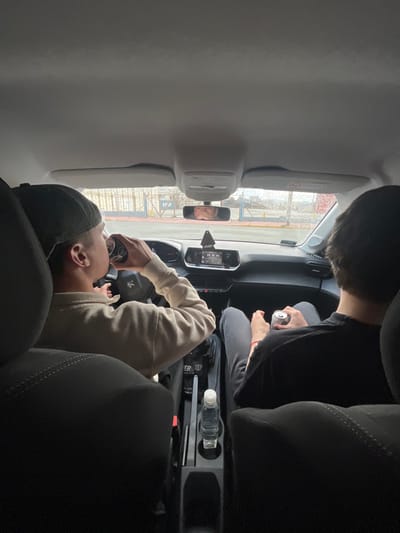 An interior shot from the rear seat of a vehicle captures two individuals in the front seats in Ushuaia, Argentina. The driver, a male wearing a dark baseball cap backward and a light-colored long-sleeved shirt, is actively drinking from a metallic can while his hands are near the steering wheel displaying a Peugeot logo. To his right, the front passenger, a male with dark hair and wearing a dark t-shirt and dark trousers, holds a similar metallic can in his left hand, resting on his lap. A clear plastic water bottle is positioned in a cup holder on the central console between the seats. Through the windshield, an exterior scene depicts a multi-colored metal barrier or fence structure (light blue/white and yellow) suggesting a port or industrial area, with visible water droplets on the glass indicating precipitation. The environment outside appears overcast.