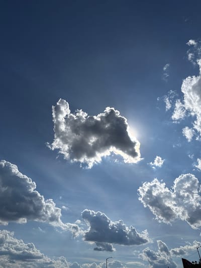 A medium shot captures a dramatic sky filled with clouds and sunlight over New York City, United States of America. The sun, partially obscured by a large, dark cloud on the left, casts rays of light through the sky, illuminating other clouds. These clouds range in color from bright white where they catch the sun to dark gray where they are in shadow, creating a stark contrast. Some of the clouds are wispy and spread out, while others are more dense and puffy. The sky itself is a deep blue, indicating a clear day despite the cloud cover. In the lower portion of the image, the tops of what appear to be streetlights and the edge of a red and black sign are visible, suggesting an urban environment. There are no people or animals clearly depicted in the image, and no explicit actions or interactions are taking place beyond the natural phenomenon of clouds and sunlight. The overall scene conveys a sense of natural beauty and the power of light.
