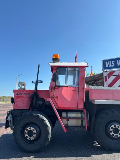 A bright red MB Trac tractor, likely a 200 series model based on the visible lettering, is parked on a brick-paved surface. The tractor has large, rugged tires, a prominent exhaust pipe, and a cab with tinted windows. A metallic elephant figurine is mounted on the left fender. A bright orange warning light sits atop the cab. Behind the tractor, several colorful flags, including red and green ones, are visible. A blue sign with white lettering that reads "VIS VAN FLOQR" is affixed to a structure on the right. The sky is clear and blue, suggesting a sunny day. The general setting appears to be an outdoor area, possibly near a coast, given the context of Zandvoort, a Dutch coastal town.
