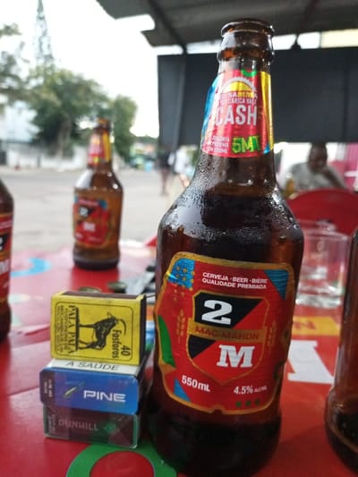 The image shows a close-up of a bottle of "2M Mac-Mahon" beer on a red table. The beer is in focus and the table is in the foreground. There are two other beer bottles out of focus in the background.  In the bottom left corner, there is a stack of three cigarette packs, one blue, one red, and one yellow. There is a box of matches with a yellow label featuring a brown goat and "Fosforos 40" written on it. The scene is likely a bar or restaurant in Maputo, Mozambique. The location is identified by the coordinates provided.  The image is likely taken by a person who is drinking beer. The image is well-lit and the colors are vivid. The lighting is coming from an overhead light source, possibly the sun or an artificial light source.  The image has a resolution of 1080 x 1920 pixels.
