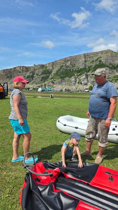 In Таш-Асты, Russia, on a grassy field with a large, rugged mountain range in the background, three people are present. A woman wearing a pink cap, grey t-shirt, and blue shorts stands on the left, facing right. A man in a camouflage cap, blue t-shirt, and camouflage shorts stands on the right, facing left. Between them, a young boy in a blue cap and a patterned t-shirt is crouched down, reaching towards a deflated red and black inflatable raft. A partially inflated white inflatable boat is positioned behind the boy and to the right of the woman. The environment suggests a recreational or preparation activity for water-based activities.