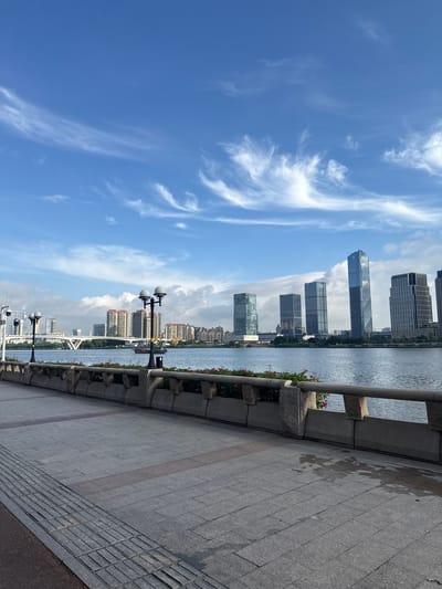 The image shows a waterfront promenade in Haizhu District, China, likely overlooking the Pearl River. In the foreground, a paved walkway with a textured tactile paving section for visually impaired individuals leads towards the right. A low stone wall with square planters filled with pink flowers separates the walkway from the river. Two ornate lampposts with globe lights stand along the promenade. Across the river, a city skyline is visible, featuring a mix of modern high-rise buildings and older residential blocks. A white arched bridge spans the river in the distance. Several boats are present on the water, including a larger vessel with a visible deck. The sky is blue with wispy white clouds. The scene depicts an urban waterfront environment.
