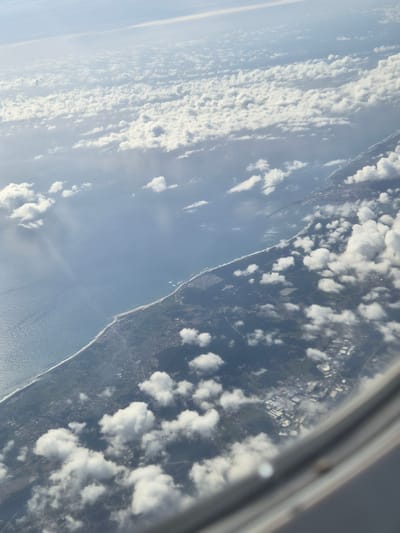 The image, taken from an airplane window, shows the coastline of Barcelos, Portugal, partially obscured by clouds. The Atlantic Ocean stretches along the left side of the frame, bordered by a sandy beach and the coastline. Beyond the coastline, the land is visible with areas of green indicating vegetation and possible structures, appearing like buildings or industrial sites. Part of the airplane's window frame is visible at the upper right corner.