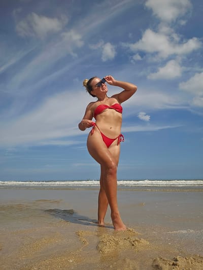 A young woman is lying on a sandy beach wearing a red bikini. She is lying on her side with her legs extended and is looking towards the camera with a smile. She is wearing sunglasses and her hair is pulled up in a bun. The beach is wet from the tide, and there are waves in the background. The sky is blue with some clouds. The time of day appears to be afternoon. The scene is set at Phu Thuy Ward, Vietnam. There is no visible text in the image.