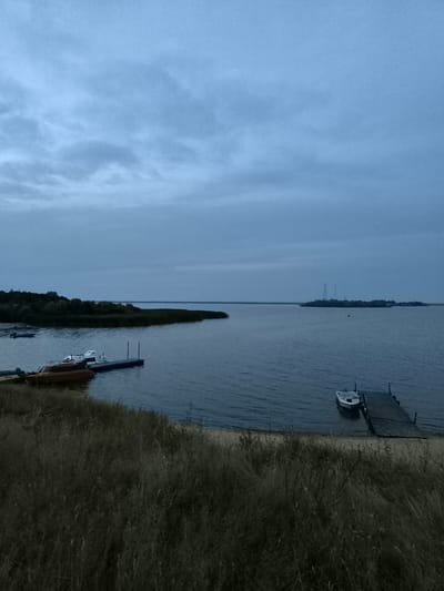 The image depicts a serene, overcast day on the water in Ostrov Im. Belova, Russia. The foreground is dominated by tall, dry grasses bordering a sandy shore. A calm, dark blue body of water stretches out, meeting a distant, tree-lined shore on the left and a low-lying island with a tall transmission tower on the right. In the middle ground, two wooden docks jut out into the water. The dock on the left has three small boats tied to it, one orange and two white. The dock on the right has a single white boat moored. The sky is a soft, muted blue with wispy, light gray clouds. The overall atmosphere is tranquil and somewhat melancholic.