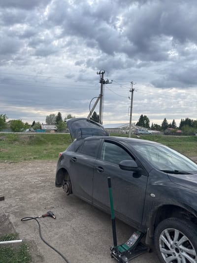 The image shows a black car with a flat tire on a dirt road in Druzhino, Russia. The car is jacked up, and the front driver-side wheel is missing. An impact wrench and lug nuts are lying on the ground near the wheel hub. The trunk of the car is open. Power lines are visible in the background. The sky is overcast. There are homes and trees in the background, indicating a residential area.