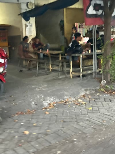 In Gelam, Indonesia, an outdoor seating area at what appears to be a food stall or small restaurant is captured in the image. Several individuals are seated at wooden tables, some engaged in conversation or eating. A red motorcycle is partially visible on the left side of the frame. The ground is paved with interlocking bricks, and scattered dry leaves suggest an outdoor setting. In the background, there are structures with awnings and a sign with graphic elements. The lighting is dim, indicating it may be evening or an overcast day.
