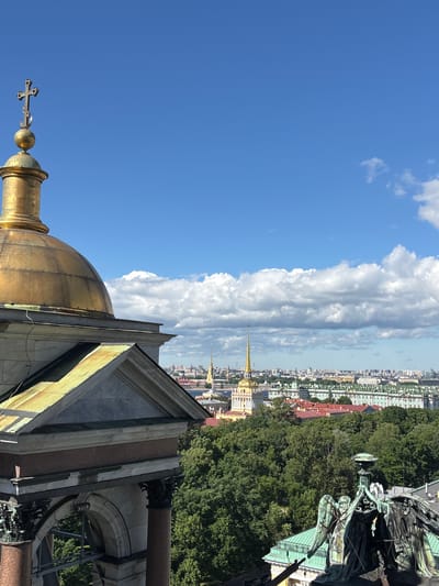 This media file is a vertical, wide-angle photograph taken from a high vantage point, offering a panoramic view of a cityscape under a bright, partly cloudy sky. The foreground features the ornate golden dome of a building, topped with a cross. Adjacent to the dome, a section of stone architecture with triangular pediments and decorative elements is visible.
The middle ground reveals a densely built urban landscape with a mix of building styles and colors, suggesting an older city. Tall, slender spires, some golden, punctuate the skyline. Lush green trees are interspersed throughout the urban fabric, creating a contrast between nature and the built environment.
In the upper right portion of the image, a large, detailed sculpture of winged figures, possibly angels, is visible, contributing to the sense of grandeur and possibly historical significance. The sculpture appears to be made of a dark, weathered metal.
The sky is a vibrant blue, with large, fluffy white clouds dominating the left side of the frame and stretching horizontally across the scene. The lighting suggests it is daytime, likely late morning or early afternoon, with the sun casting distinct shadows.
The overall impression is one of an expansive, historic European city, viewed from a significant height, perhaps from the top of a cathedral or tower. The weather appears pleasant, with clear skies and some clouds adding visual interest. There is no visible text in the image.