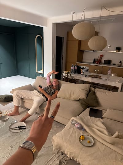 The image depicts an indoor scene, likely a modern apartment in Nice, France, with a living room and kitchen area visible. In the foreground, a person's hand wearing a watch and rings is extending towards the camera, making a peace sign. In the background, a person is reclining on a large, L-shaped sofa, holding a mobile phone and also making a peace sign with their other hand. The sofa is covered in light-colored cushions and throws, with a glass coffee table in front of it holding plates, knives, and a small bowl.
The kitchen area features light wood cabinetry, a countertop with various bottles of water and other items, and a stove. Two large, spherical paper lanterns hang from the ceiling in the kitchen space. To the left, a dark green wall with built-in cabinets and a tall, oval mirror is visible, suggesting a stylish and contemporary interior design. The overall lighting is soft and ambient.