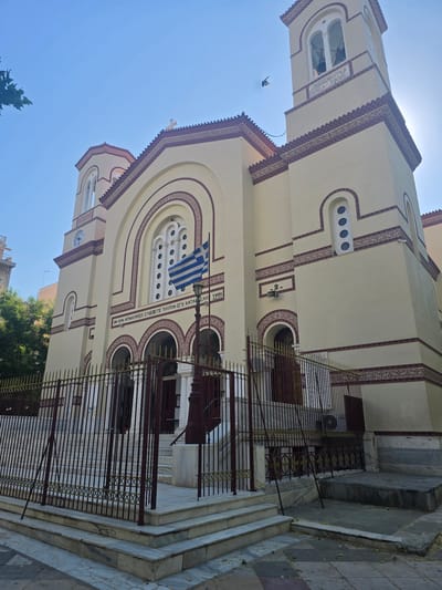 The image shows the Church of Agios Georgios Karytsi in Athens, Greece, under a clear blue sky. The church is a cream-colored building with red decorative trim around its windows, arches, and roofline. It features two bell towers, a large arched central entrance with a Greek flag on a flagpole, and a fenced-off area with marble steps leading to the entrance. The fence is made of dark metal bars with a gold decorative top. There are some trees to the left of the church and a glimpse of a modern building in the background. A bird is seen flying in the sky. The overall scene depicts a sunny day in front of a prominent religious landmark in Athens.