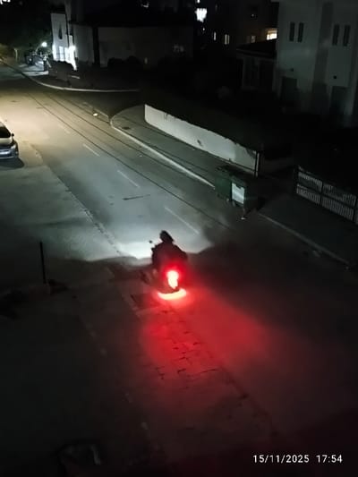 At night, a person on a motorcycle is driving down a street. The motorcycle's headlights are illuminating the road ahead, and the taillight is casting a red glow behind it. The street is lined with buildings and trees, and the sky is dark. The timestamp reads "15/11/2025 17:54" at the bottom right.