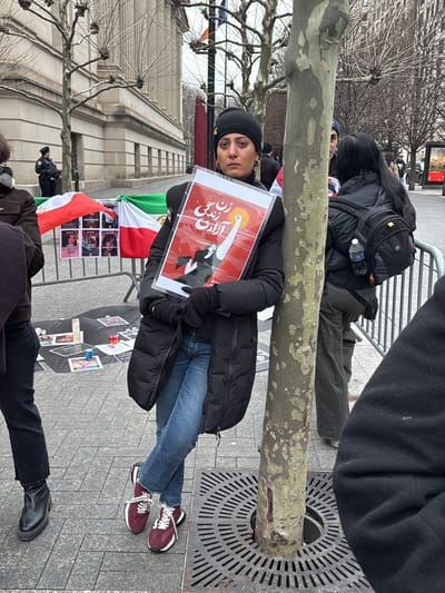 This image captures an outdoor scene in Midtown Manhattan, New York City, likely near the United Nations Headquarters area, during what appears to be a protest or vigil.

In the foreground, a woman of Middle Eastern descent stands, leaning her left side against the trunk of a bare deciduous tree. She wears a black knit beanie, a black puffer coat, blue jeans, and maroon sneakers with white accents. Her expression is somber, with apparent tear stains on her cheeks. She holds a framed poster with a red background, featuring white Persian script at the top ("زن زندگی آزادی" - Woman, Life, Freedom) and a stylized illustration of a female figure in black raising a white, radiating fist. The tree is surrounded by a circular metal grate embedded in the paved sidewalk.

To the far left, partially visible, is a person in dark clothing and black boots. In the mid-ground, behind a metal crowd control barrier, several Iranian flags (green, white, and red stripes, notably missing the central emblem of the Islamic Republic) are draped. On the paved ground behind these flags, a makeshift display includes black cloths, numerous small photographs (possibly of victims), several lit candles (some red, some white), and small plastic containers, arranged in what appears to be a memorial.

Further in the background, to the left, a uniformed police officer in a dark coat and hat stands near a large, ornate classical stone building with multiple windows and columns. To the right, another individual with long dark hair, wearing a black puffer coat and a black backpack (with a plastic water bottle in a mesh pocket), stands behind the metal barrier. The sky is overcast, and the bare trees suggest a winter or early spring setting. The ground is paved with rectangular and square stone tiles.