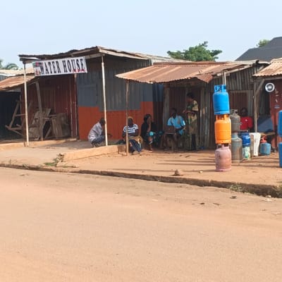 This image depicts a street scene in Rido, Nigeria. A row of makeshift structures with corrugated metal roofs lines a dirt road. A sign above one of the structures reads "WATER HOUSE." In front of these structures, several people are gathered.
On the left side, a man in a striped shirt and jeans squats near a white pole. Next to him, three people are seated: a woman in a striped shirt and blue skirt, a woman in a black headscarf and patterned skirt, and a young man in a blue t-shirt who is using a laptop. Further to the right, a woman in a patterned dress stands, possibly interacting with the man using the laptop.
In the foreground, stacked gas cylinders are visible, with a blue one at the top, an orange one in the middle, and a reddish-brown one at the bottom. Other gas cylinders and various objects, including a red fire extinguisher, are also present near the structures. The environment appears dusty and sun-drenched, with sparse vegetation growing along the roadside. The overall impression is that of a local marketplace or service area.