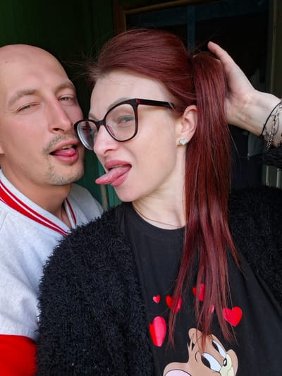 A close-up, eye-level, medium shot shows a bald man with a stubble beard and a woman with long, red hair and glasses. The man is on the left, his head tilted, his right eye winking, and his tongue sticking out slightly. The woman is on the right, her head tilted to the left, her tongue extended. They are both wearing casual clothing: the man a grey sweatshirt with red and white stripes on the collar, and the woman a black fuzzy cardigan over a black t-shirt with red hearts and a cartoon character print. The background is dark and appears to be an outdoor setting with a wooden door frame, suggesting a rural or suburban environment. The lighting is natural, illuminating their faces.