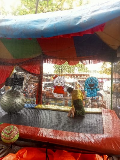 A child is kneeling on a black trampoline within a colorful, netted enclosure. A large, textured ball sits to the left, and a colorful ball rests on the red padded edge. Cutout figures of Hello Kitty and Doraemon hang inside the netting. The enclosure has a striped tent roof in shades of blue, red, and green. A blurred background shows hints of trees and outdoor structures.