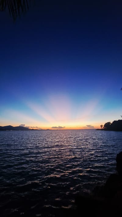 This is a landscape photograph of a sunset over the ocean at Baan Plai Laem, Thailand. The image is oriented vertically. The sky transitions from a deep blue on the left to a soft orange and yellow near the horizon on the right. Sun rays emanate from a point just above the horizon, creating a dramatic effect. Clouds are scattered across the sky. The ocean occupies the right half of the image, with textured waves reflecting the colors of the sunset. In the distance, the silhouette of land is visible along the horizon, suggesting a coastline or islands. The foreground on the far left shows a dark silhouette of palm fronds, adding a tropical element to the scene. The overall mood is serene and peaceful, characteristic of a sunset. There is no visible text in the image.