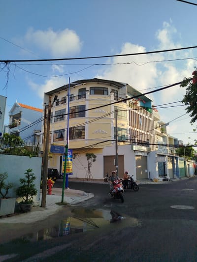 The image depicts a street scene in Ho Chi Minh City, Vietnam. The sky is blue with scattered white clouds. Overhead, there are several electrical wires crisscrossing the sky. The corner of a building with a distinct yellow facade and ornate trim is visible, with a green-and-blue street sign indicating "YÊN BÁI." A red fire hydrant is situated near the sign. Two people on motorbikes are driving on the asphalt road, which has a puddle reflecting the surroundings. The road also shows wear and tear. A variety of buildings are seen along the street, with greenery visible on the left side.
