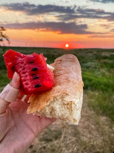 A close-up shot shows a hand holding a piece of watermelon and a section of bread against a sunset backdrop. The watermelon is red with visible black seeds and appears to have been bitten into. The bread is a pale crusty loaf. The hand has a bandage on one finger. The background features a vibrant orange and pink sunset with clouds, and a green field extending to the horizon. The scene is set outdoors in Harmanli, Bulgaria.
