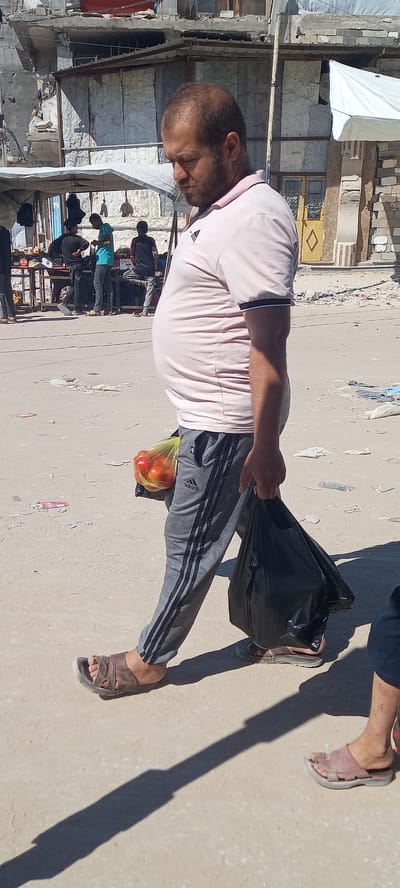 In Khan Yunis, Palestinian Territories, a man with a large belly walks from right to left, his body angled slightly away from the camera. He is wearing a light pink polo shirt with a dark trim on the collar and sleeves, and grey athletic pants with black stripes down the legs. He carries a yellow plastic bag containing red tomatoes in his left hand, and a black plastic bag in his right hand. He is wearing brown leather sandals.
In the background, other individuals are visible near a market stall. One person wears a turquoise shirt and dark pants. Another person in the background wears a dark t-shirt and pants. The environment appears to be an outdoor marketplace with some structures in disrepair, including damaged buildings with exposed rebar and concrete, and makeshift shelters with corrugated metal and fabric coverings. Debris is scattered on the sandy ground. The lighting suggests it is daytime, with strong shadows cast on the ground.