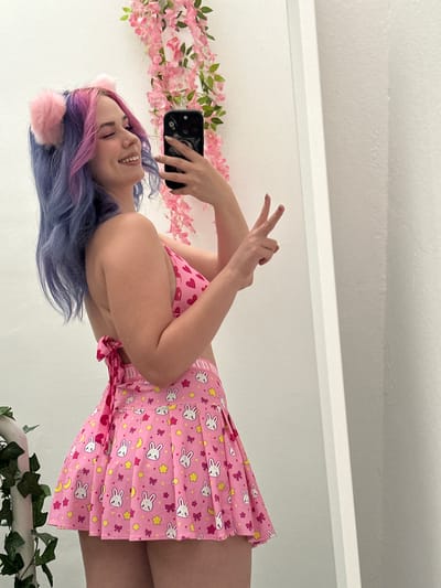 A woman with long, dyed blue and purple hair, adorned with pink cat ears, is smiling and holding a smartphone in her right hand. She's wearing a pink outfit patterned with bunnies, hearts, stars, and moons. A string of pink flowers is visible in the background on the left, while green foliage is visible on the right, likely attached to a white object. The location is likely indoors due to the plain white walls. There is no indication this is in City of Zagreb, Croatia.