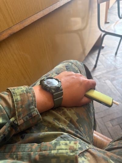 A person in camouflage clothing sits at a light brown table. They are holding a yellow electronic cigarette device. The person is wearing a green digital watch on their left wrist. The background includes a chair and wood floor. No information suggests this is located in Dolna Mitropolia, Bulgaria.