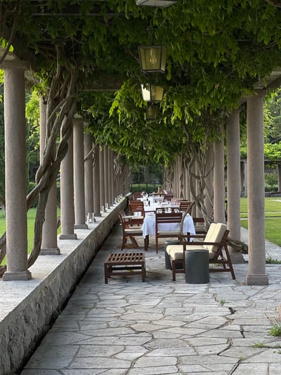 This is a full, eye-level shot of an outdoor dining area at the Aman Sveti Stefan resort in Montenegro, taken in daylight. The scene is characterized by a long, covered walkway supported by a series of thick, stone pillars. Twisted vines and lush green foliage drape over the top of the walkway, creating a natural canopy.

The walkway features a stone floor and a low, stone wall along its left side. On the right side, dining tables are set with white tablecloths and glassware, arranged to face a grassy area. There are wooden chairs with light-colored cushions, and some low wooden tables. In the foreground, two lounge chairs with cushions are visible, accompanied by a dark cylindrical stool. A row of outdoor lamps, enclosed in glass and metal casings, hang from the overhead structure, illuminating the dining area.

The background shows more greenery, including trees and manicured grass, suggesting a well-maintained outdoor space. The overall impression is one of an elegant and tranquil al fresco dining setting within the resort.