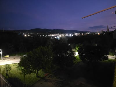 This is a nighttime long-exposure photograph taken from an elevated position, likely a balcony or window, overlooking a town or city. The sky is a deep twilight blue. In the foreground, a dark grassy area with several trees is illuminated by artificial lights. A fence runs along the left side of this area. Further into the scene, a road is visible with a yellow car parked on it, and a street lamp. Beyond the immediate foreground, a townscape is spread out, with numerous lights indicating buildings and infrastructure. In the distance, hills or mountains form a dark silhouette against the sky. Some industrial structures, possibly including power plant chimneys, are identifiable amongst the distant lights. The overall impression is of a quiet, urban or semi-urban environment at dusk or early night. The photograph was taken in the city of Pernik, Bulgaria.