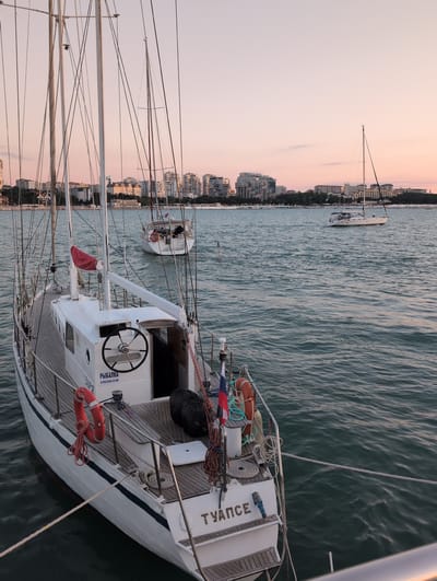 The image shows several sailboats docked in a harbor with a city skyline in the background. The sky is a soft pink and orange, indicating either sunrise or sunset. The water is calm and reflects the sky. The sailboats are white with wooden decks and masts. One sailboat in the foreground has the name "ТУАПСЕ" written on its stern. The city skyline features numerous buildings, some appearing to be apartment complexes and others with a more traditional architectural style. There are also trees lining the coast. The overall scene is peaceful and serene. Based on the text on the boats and the architectural style, the location is likely Gelendzhik, Russia.