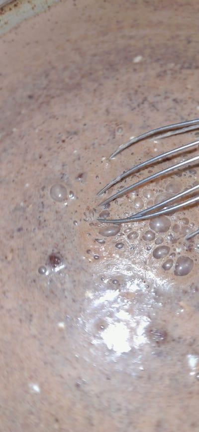 The image displays a close-up view of a light brown, frothy liquid, likely a beverage or batter, within a container that appears to be partially visible along the top edge. The surface is covered in small bubbles, with a few larger, more distinct bubbles scattered throughout. A silver whisk is submerged in the liquid on the right side of the frame, with its metal loops creating ripples and further contributing to the frothy texture. The lighting suggests a bright environment, with reflections visible on the surface of the liquid, particularly in the lower half of the image. The overall scene is domestic and focused on food preparation. There are no indications of the city of Vallecito, Venezuela, or any elements that would place the scene within a specific geographical or urban context.