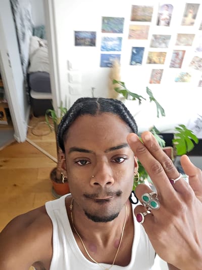 A person with dark skin, braided hair, a nose piercing, and wearing a white tank top and gold jewelry is posing for a selfie. They are wearing rings on their fingers. The background includes a wooden wall or floor and a white wall with a collection of small images or postcards displayed on it. A potted plant is also visible. The location in Pantin, France, cannot be determined from the image.