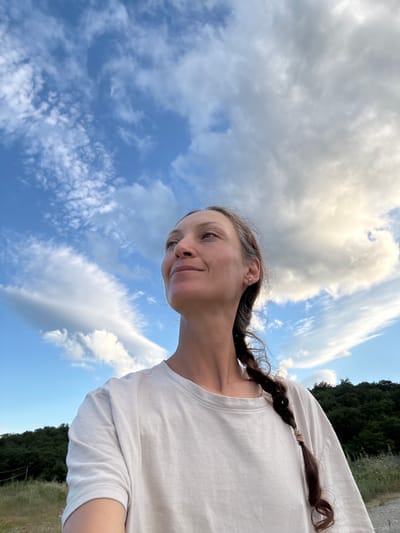 A woman is pictured from the chest up, looking to her left and slightly upwards at a cloudy sky. She has long, dark brown hair in a braid that falls over her shoulder. She is wearing a light-colored t-shirt. The background consists of a bright blue sky with scattered white and gray clouds, and a distant line of dark green trees. The photograph appears to have been taken outdoors in a natural setting.