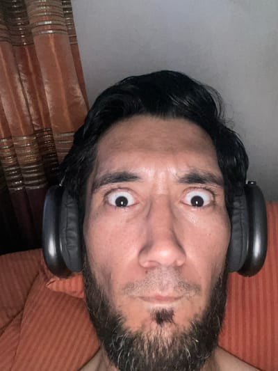 A man with black hair, a beard, and a soul patch is wearing black over-ear headphones. He is looking directly at the camera with wide eyes. He is sitting in front of a textured orange curtain and an orange couch. The image appears to be taken indoors, likely in a residential setting. There is no indication of the city of Mashrab, Uzbekistan, in the image.