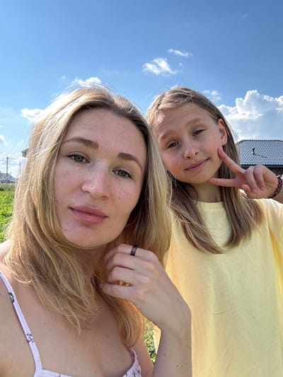 A selfie taken outdoors on a sunny day shows two individuals, likely an adult and a child, in the city of Mokino, Russia. The background features a clear blue sky with scattered white clouds, green grass, and the rooflines of houses, indicating a residential area.
On the left, closer to the camera, is an adult with blonde hair, green eyes, and fair skin. She is wearing a light-colored top with a floral pattern. Her hair is styled loosely, framing her face. She is looking directly at the camera.
To the right of the adult is a younger person, also with blonde hair and fair skin. This individual is wearing a yellow t-shirt and is making a peace sign with their fingers near their face. They are looking towards the camera with a slight smile.
The lighting suggests it is daytime, with the sun casting light on the subjects. The overall scene conveys a casual, happy moment captured outdoors.