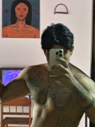 The image captures a mirror selfie of a shirtless individual in an indoor setting, possibly in Guatacaral, Venezuela. The subject, a person with dark, wavy hair, holds a light-colored smartphone (specifically an iPhone with a triple-lens camera array) in their right hand, obscuring the majority of their face. Extensive dark tattoos are visible across the individual's chest and both arms, including a large design on the chest and a solid dark band on the left forearm.
The background, visible in the mirror's reflection, consists of a light-colored wall. Above and to the left of the subject's head, a rectangular painting is mounted, depicting the upper torso of a person with dark hair, green eyes, and orange-toned skin against a dark blue background with abstract, possibly leaf-like, elements. To the right of the painting, a small, curved, brown D-shaped object is affixed to the wall. In the lower-left quadrant of the image, a segment of a wooden furniture item with horizontal slats is visible. Below the painting and adjacent to the subject's left side, a screen (likely a television or monitor) displays a vibrant blue and purple illuminated graphic.