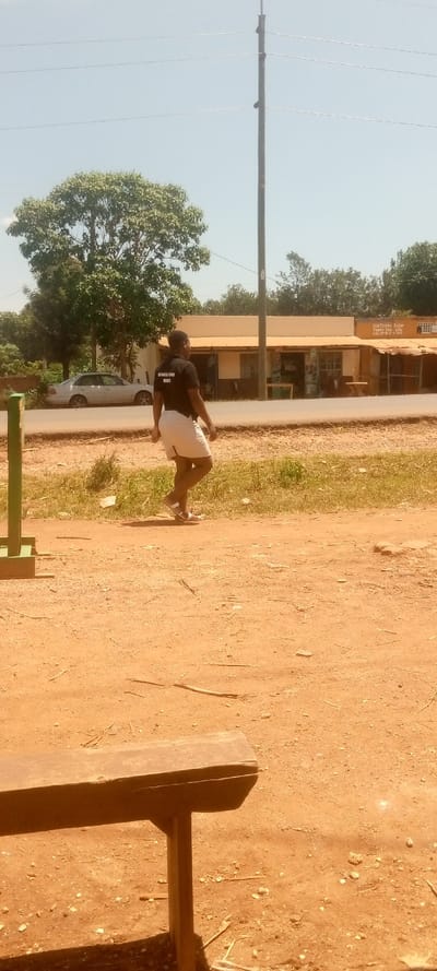 The image shows a man walking on a dirt road in Kivaywa, Kenya. The man is wearing a black t-shirt with a white inscription and white shorts. He is walking away from the camera and towards a small building with a sign that reads "Startimes Solar Products and Services. Call 0700 22 222." There is a wooden bench in the foreground. The man is walking on a dirt road that is surrounded by grass and trees. The sky is clear and blue. The location is latitude 0.56, longitude 34.806 in Kivaywa, Kenya.