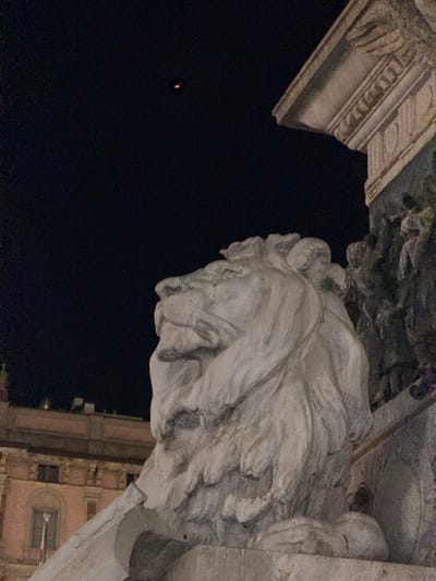 The image depicts a nighttime scene in Milan, Italy, featuring a close-up view of a large stone lion sculpture. The sculpture is positioned in the foreground and faces left, with its head and mane being the dominant elements. The lion appears to be part of a larger monument, with ornate stonework visible to the right. In the background, a building with a pink facade and arched windows is illuminated, suggesting an urban environment. Above the building, a dark sky is visible, with a faint, distant light source appearing as a small, red and yellow speck. The overall impression is of a nocturnal, monumental setting in Milan.