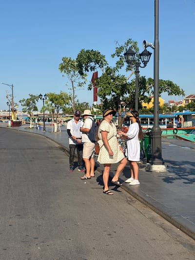 In Hội An Ward, Vietnam, on a bright, sunny day, four individuals are gathered on a paved walkway adjacent to a street. A man in a white polo shirt and dark pants, wearing a baseball cap, stands facing a person in a light-colored short-sleeved shirt and shorts, also with a hat. To the right, two women are interacting. One woman wears a short, yellow dress with a floral pattern and a straw hat, while the other is dressed in a short, white dress. They are positioned near a lamppost that also has a speaker attached. In the background, a line of colorful boats is docked along a body of water, and various buildings, some with yellow facades, are visible. The environment suggests a bustling tourist area.
